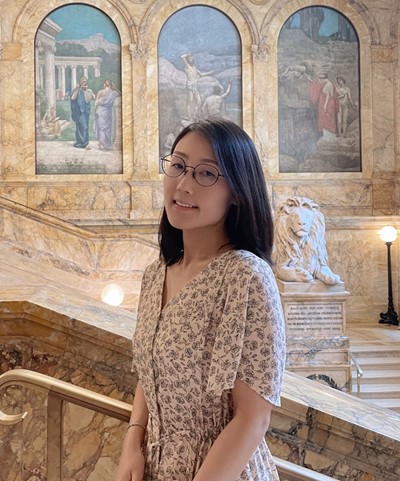 Anna from the torso up smiling wearing a beige floral dress against marble infrastructure 