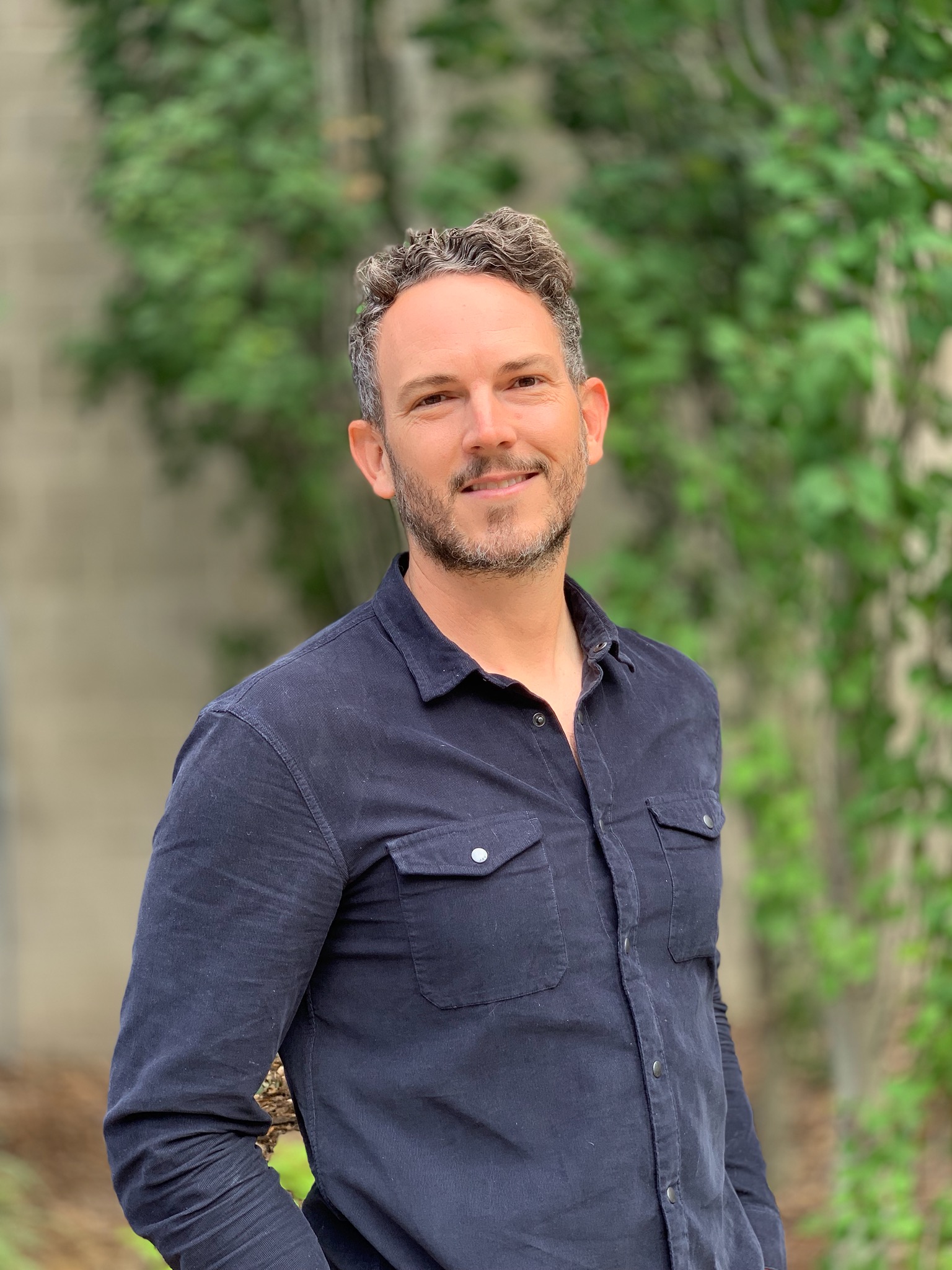 A photo of Avery from the torso up - he is wearing a gray button-up against a blurred nature background
