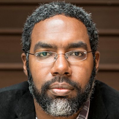 Close up headshot of Deen, straight-faced with a black popped collar against a brown backdrop 
