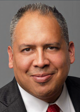 Dr. Valeriano smiling wearing a suit. Photo from the shoulders up and against a plain gray background.