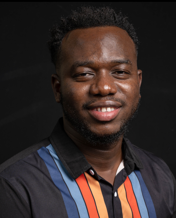Eugene from the shoulders up smiling and wearing a black striped button-up against a black background
