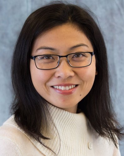Ke/"Maddie" from the shoulders up smiling and wearing a white turtle neck against a light gray background