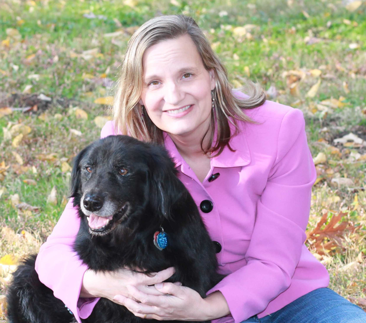 Jana smiling in a pink blasier and hugging her Black dog against a grassy background.