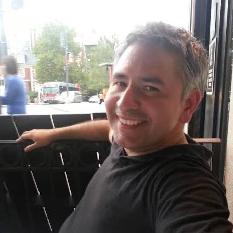 Man with short grey hair sitting casually at a restaraunt, smiling at the camera