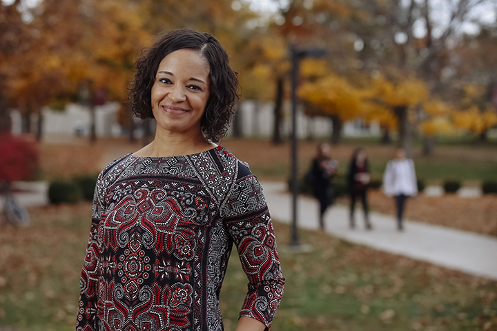 Rachel from the waist up wearing a black and red patterned shirt with her hand on her hip smiling against a fall landscape