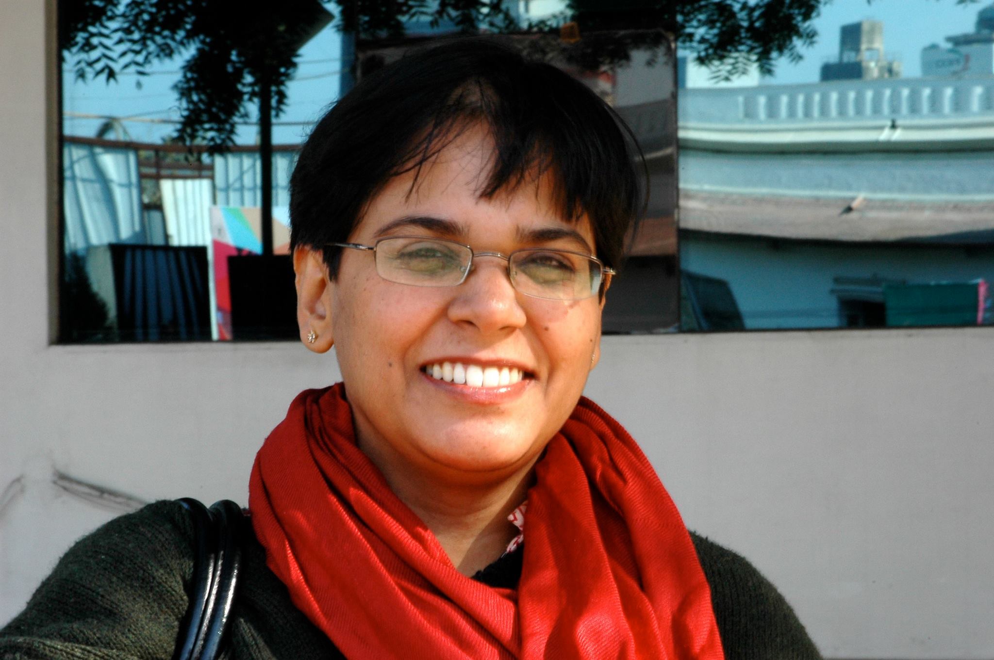 Sananda from the shoulders up smiling and wearing a black shirt and red scarf against a white wall with photos landscape hung on it
