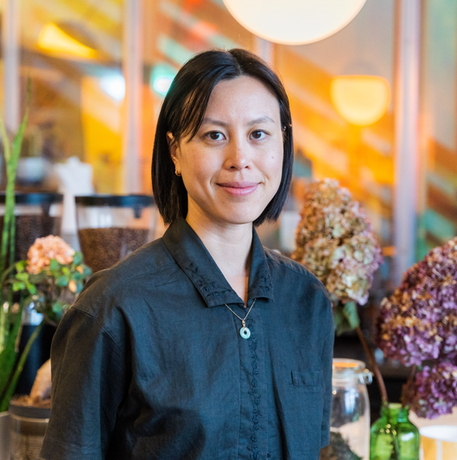 Sarah smiling wearing a dark button up shirt against a blurred indoor background with plants, coffee, and lights.