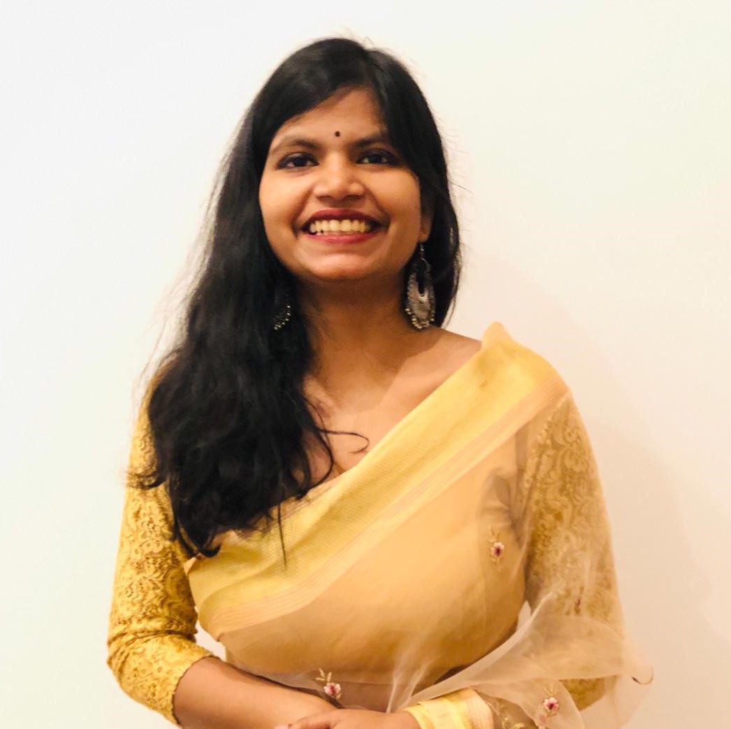 Sneha from the torso up similing and wearing a yellow dress/saree against a white backdrop/wall