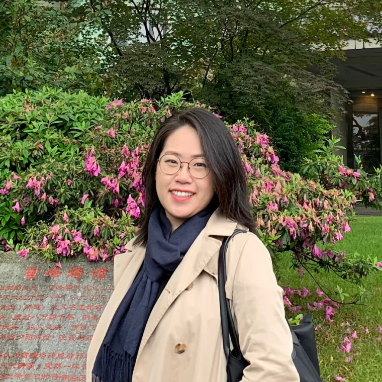 Yu from the torso up smiling wearing a black shirt and beige jacket against a green space background